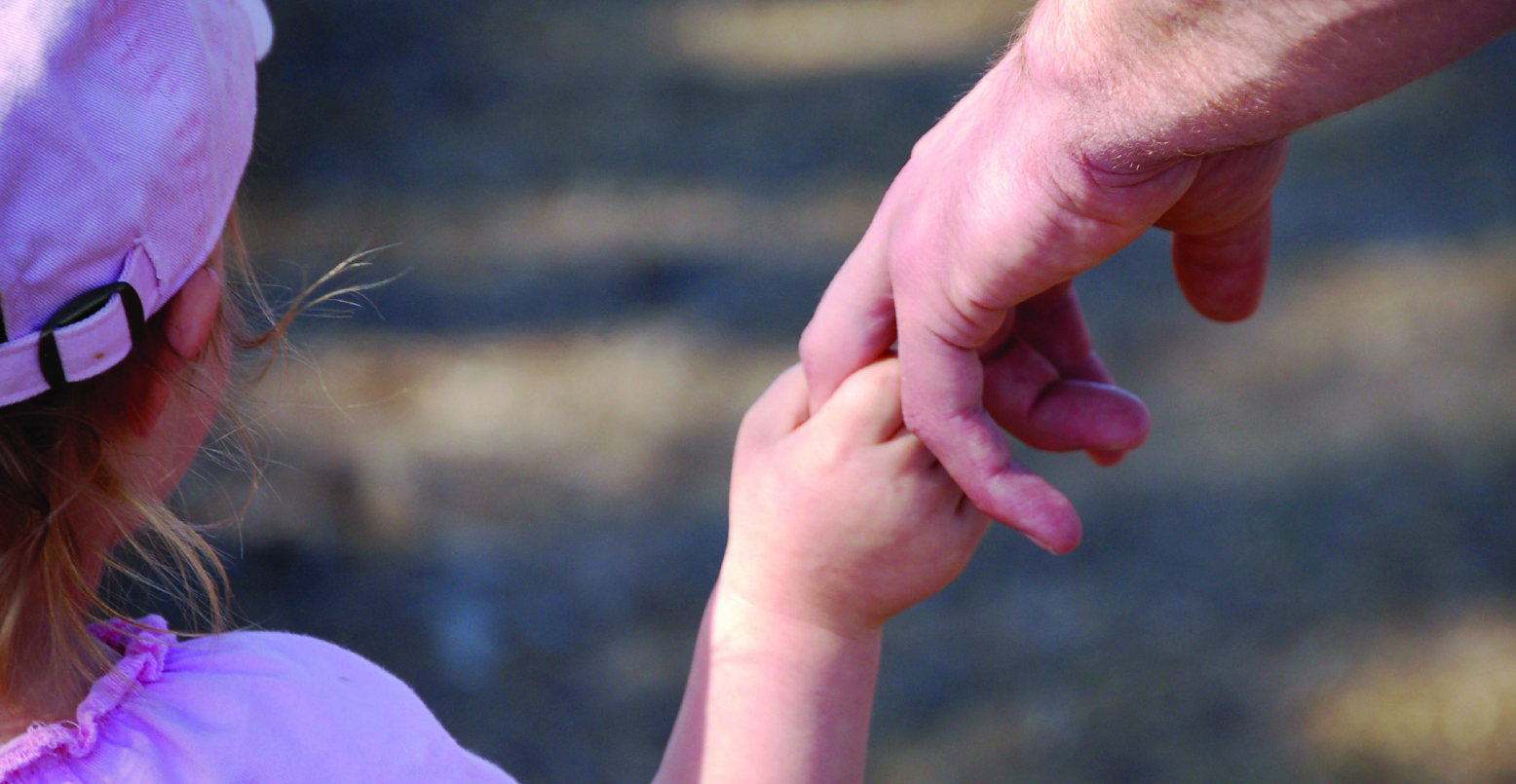 Viewed from behind, a young child, in a pink hat and top, holding an adult's hand.